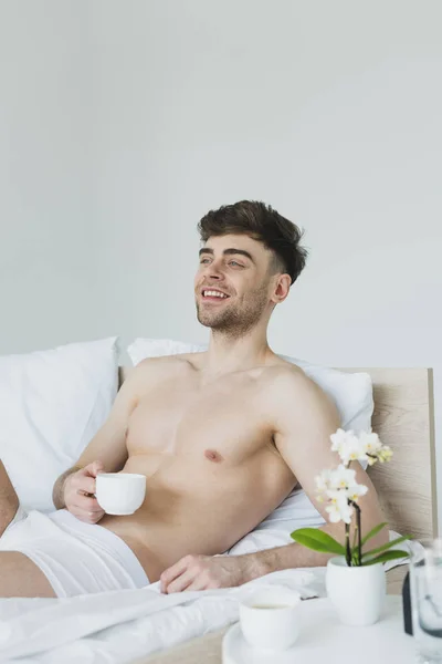 Homem alegre em roupa interior segurando xícara de café enquanto deitado na cama e olhando para longe — Fotografia de Stock