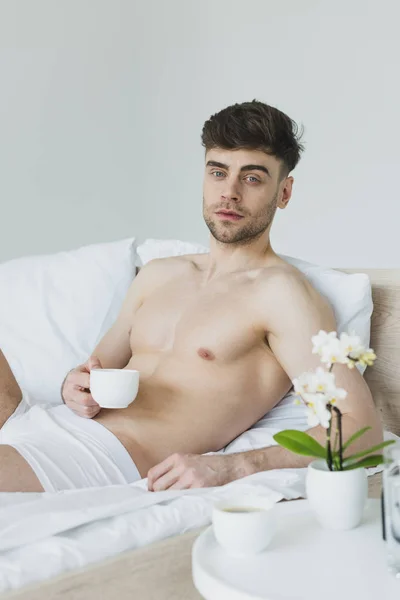 Pensive man in underwear holding coffee cup while lying in bed and looking at camera — Stock Photo