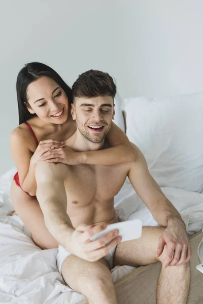 Feliz pareja interracial en ropa interior sentado en ropa de cama blanca y tomar selfie con teléfono inteligente - foto de stock