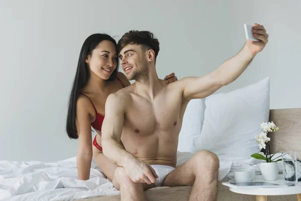 Heureux couple interracial assis sur la literie blanche en sous-vêtements et de prendre selfie avec smartphone — Photo de stock