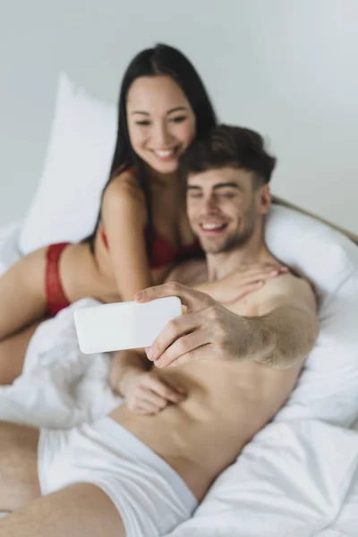 Избирательный фокус счастливой межрасовой пары в нижнем белье, лежащей в постели и делающей селфи со смартфоном — стоковое фото