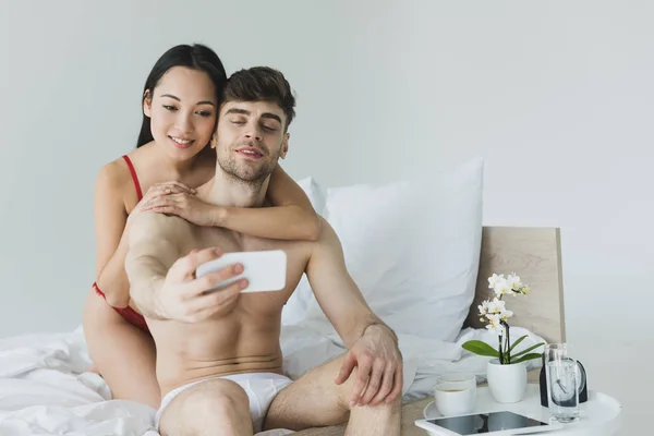 Feliz casal interracial sentado em roupa de cama branca em roupa interior e tomando selfie com smartphone — Fotografia de Stock