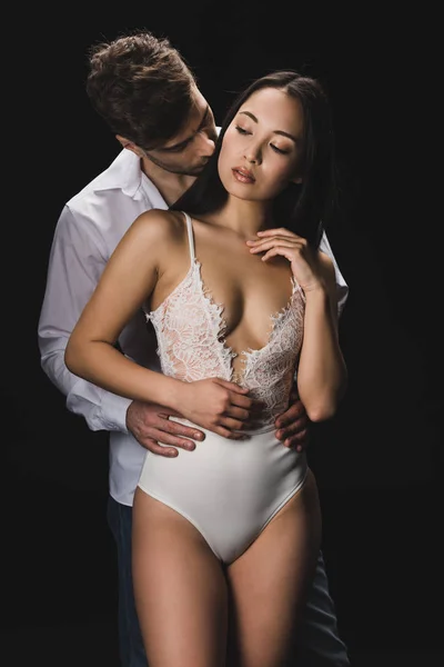 Guapo joven abrazando y besando atractivo asiático novia en blanco lencería aislado en negro - foto de stock