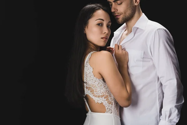 Attractive asian woman in white lingerie standing near boyfriend and looking at camera isolated on black — Stock Photo