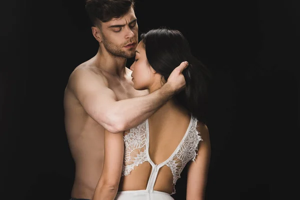 Guapo sin camisa hombre abrazando sexy novia en blanco lencería aislado en negro - foto de stock