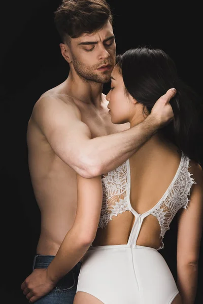 Joven sin camisa hombre abrazando sexy novia en blanco lencería aislado en negro - foto de stock