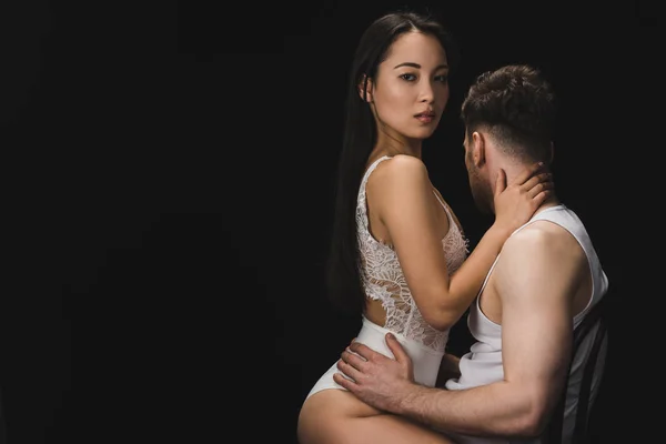 Sexy asiático mujer en blanco lencería sentado en rodillas de novio y mirando la cámara aislado en negro - foto de stock