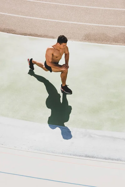 Mixed race shirtless sportsman in sneakers stretching at stadium — Stock Photo