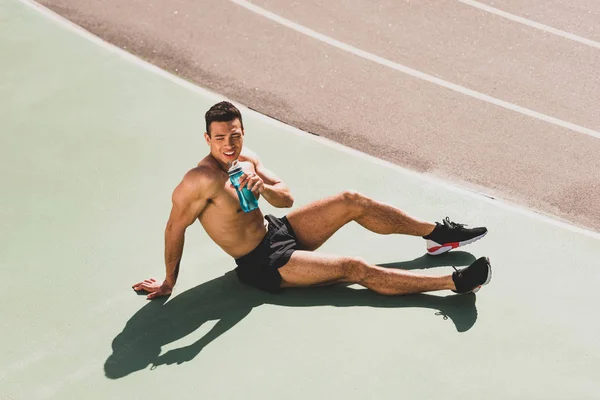 Sonriente deportista de carrera mixta sentado en el estadio y sosteniendo la botella de agua - foto de stock