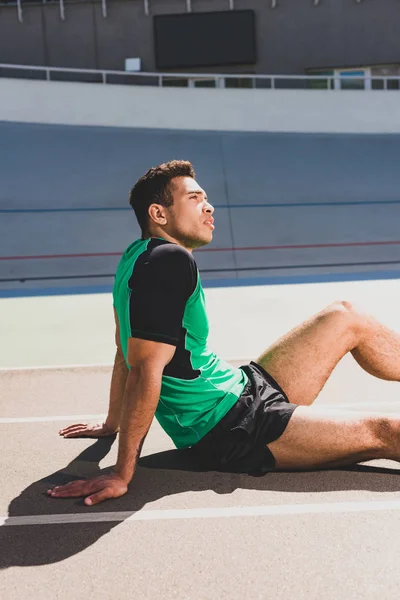 Уставший двурасовый спортсмен в спортивной одежде, сидящий на стадионе — стоковое фото