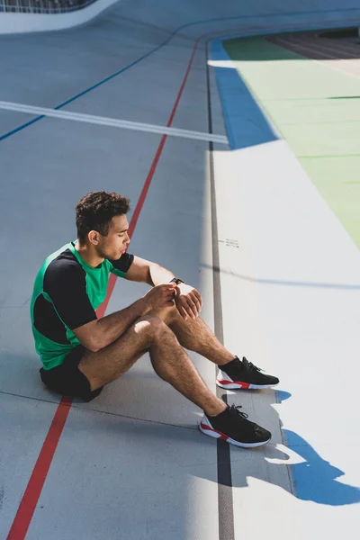 Apuesto deportista de carreras mixtas sentado en pista de atletismo en el estadio y mirando smartwatch - foto de stock