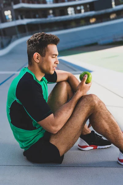 Vue latérale du sportif de course mixte assis sur la piste de course et regardant la pomme verte — Photo de stock