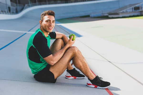 Deportista de carrera mixta sentado en pista de atletismo en el estadio, mirando a la cámara y sosteniendo manzana verde - foto de stock