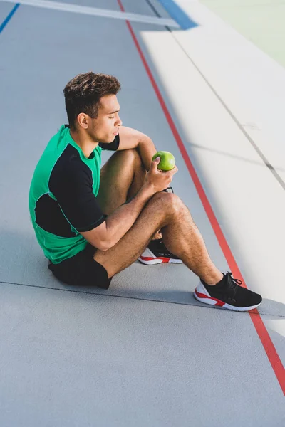 Desportista de raça mista sentado na pista de corrida e segurando maçã verde — Fotografia de Stock