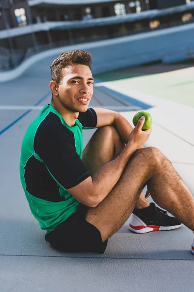 Schöner Mischlingssportler sitzt auf der Laufbahn, blickt in die Kamera und hält grünen Apfel in der Hand — Stockfoto