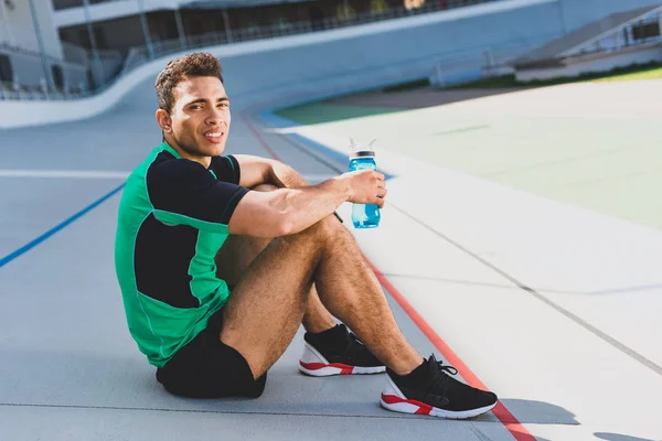 Mixed Race Sportler sitzt auf Laufbahn im Stadion, blickt in die Kamera und hält Flasche mit Wasser — Stockfoto