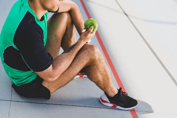 Обрезанный вид спортсмена, сидящего на беговой дорожке и держащего зеленое яблоко — стоковое фото