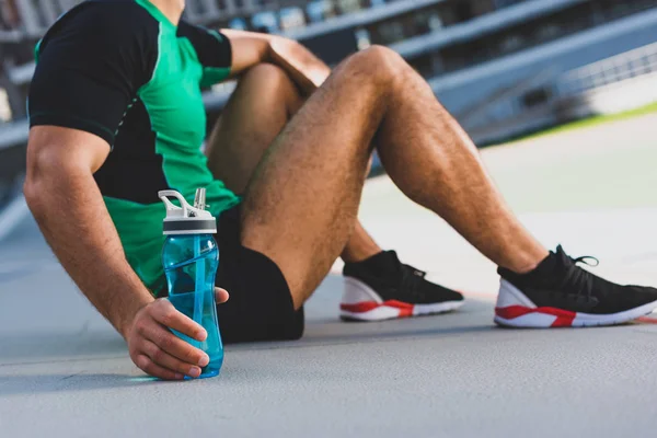 Ausgeschnittene Ansicht eines Sportlers, der auf der Laufbahn sitzt und Flasche mit Wasser in der Hand hält — Stockfoto