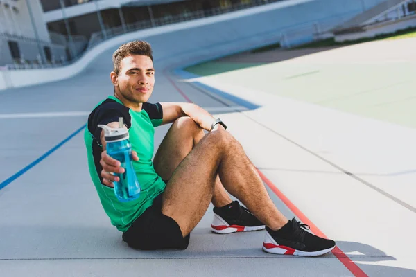 Deportista de carrera mixta sentado en pista de atletismo en el estadio, mirando y presentando botella con agua en la cámara - foto de stock