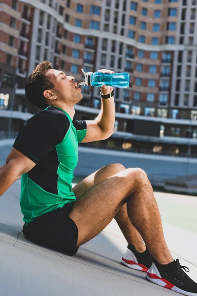 Mixed race sportsman sitting on running track at stadium and drinking water from blue bottle — Stock Photo