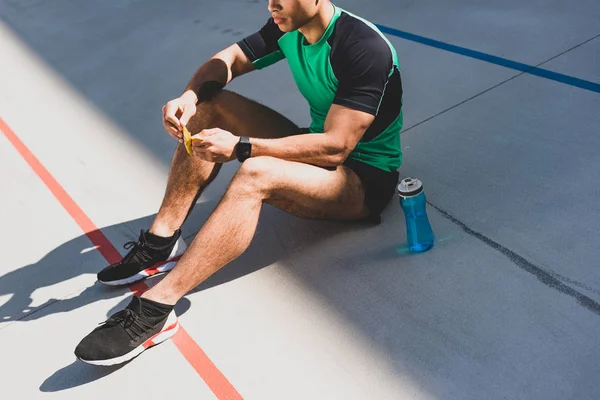 Cropped view of sportsman sitting on running track near blue bottle and opening chocolate bar — Stock Photo