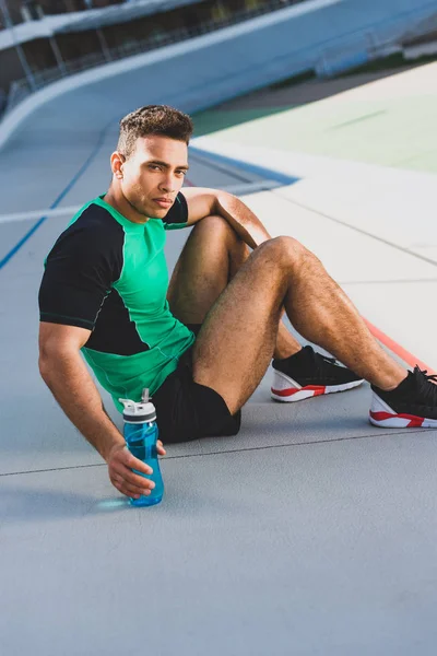 Sportif mixte assis sur la piste de course, regardant sérieusement la caméra et tenant bouteille avec de l'eau — Photo de stock