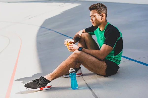 Seitenansicht eines Mixed Race Sportlers, der auf der Laufstrecke in der Nähe einer blauen Flasche sitzt und eine Schokoriegel öffnet — Stockfoto