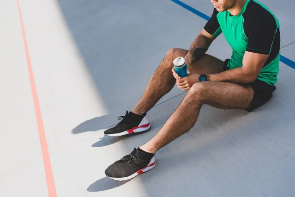 Vista cortada de desportista sentado na pista de corrida e segurando garrafa azul com água — Fotografia de Stock