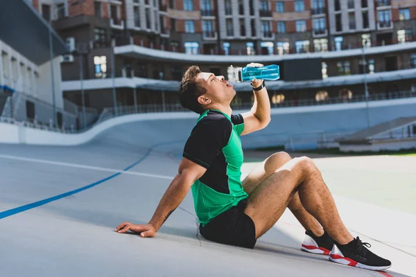 Вид збоку змішаної гонки спортсмен сидить на біговій доріжці на стадіоні і п'є воду з блакитної пляшки — стокове фото