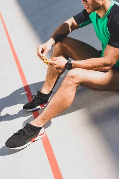 Cropped view of sportsman sitting on running track and opening chocolate bar — Stock Photo
