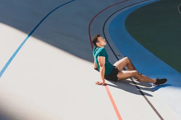 Vista lateral do desportista de corrida mista sentado na pista de corrida no estádio em tempo ensolarado — Fotografia de Stock