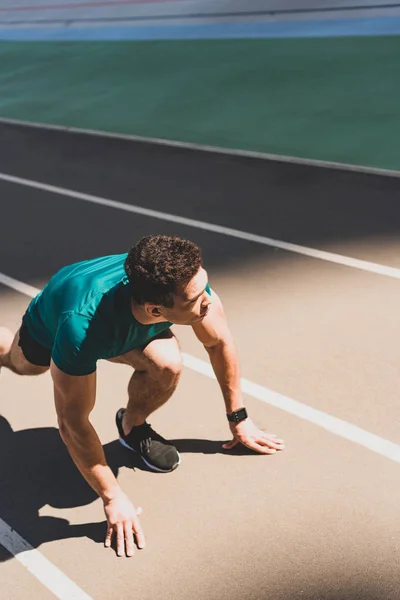 Atleta di razza mista in posizione di partenza su pista da corsa, guardando lontano allo stadio — Foto stock