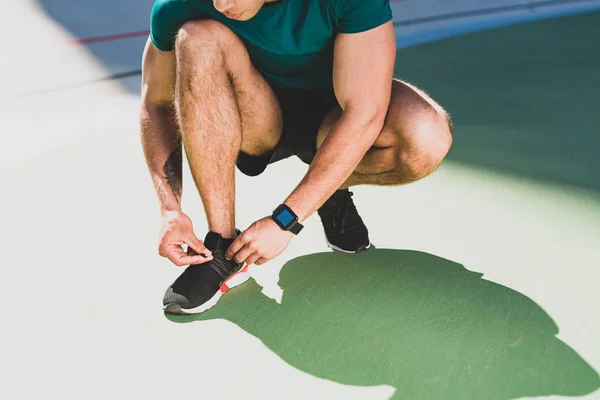 Обрезанный вид спортсмена, шнуряющего кроссовки, стоящего на зеленом полу — стоковое фото