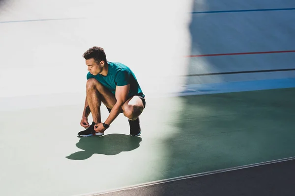 Mixed race sportsman lacing up sneakers at stadium, standing on green floor — Stock Photo