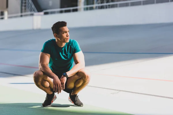 Frontansicht eines Mixed-Race-Sportlers, der im Sonnenlicht im Stadion hockt und wegschaut — Stockfoto