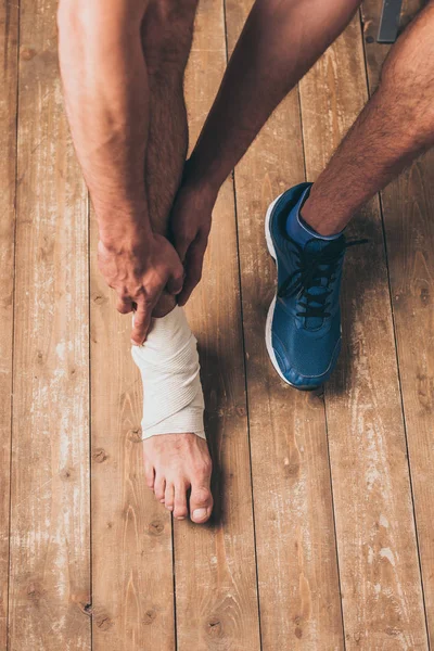Tiro cortado de desportista sentado em um tênis e tocando a perna ferida em bandagem elástica — Fotografia de Stock
