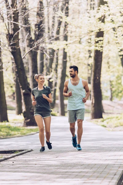 Joven hombre y mujer en ropa deportiva sonriendo mientras trota en el parque verde - foto de stock