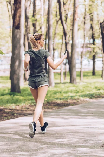Vue arrière de la jeune femme tenant smartphone et écoutant de la musique dans les écouteurs tout en faisant du jogging dans le parc — Photo de stock