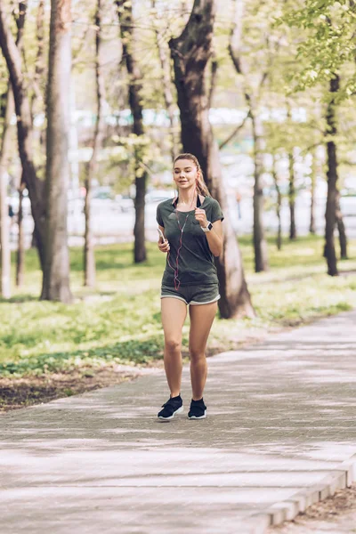 Graziosa giovane donna che tiene smartphone e ascolta musica in auricolare mentre corre nel parco — Foto stock