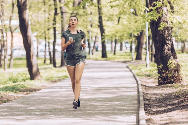 Attractive young woman holding smartphone and listening music in earphones while jogging in park — Stock Photo