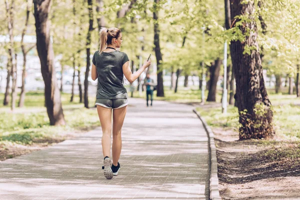 Back view of young sportswoman jogging in park while holding smartphone and listening music in earphones — Stock Photo