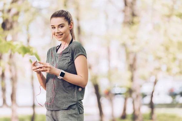 Jolie femme en vêtements de sport souriant à la caméra tout en tenant smartphone et écouter de la musique dans les écouteurs — Photo de stock