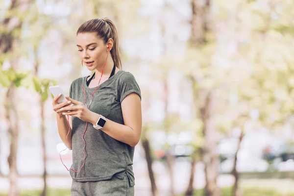 Beautiful sportswoman holding smartphone and listening music in earphones — Stock Photo