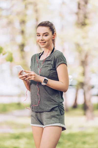 Attractive woman in sportswear using smartphone and listening music in earphones while smiling at camera — Stock Photo