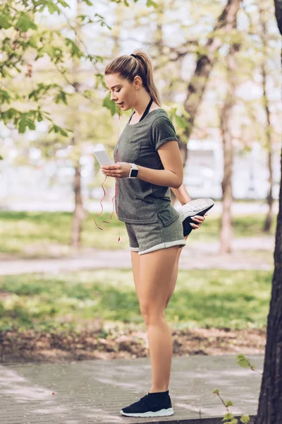 Pretty sportswoman stretching in park while using smartphone and listening music in earphones — Stock Photo