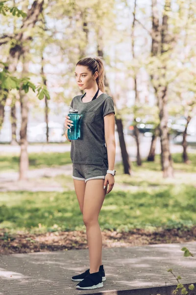 Hübsche Frau in Sportbekleidung hält Sportflasche in der Hand und schaut im Park weg — Stockfoto