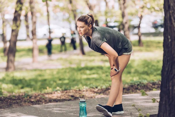 Pretty sportswoman touching injured knee while standing in park near sport bottle — Stock Photo