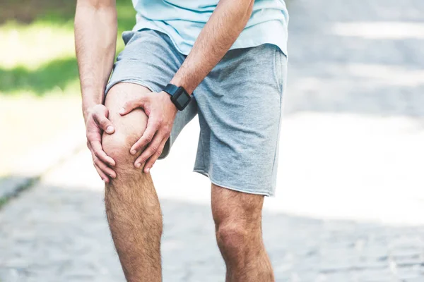 Partial view of sportsman in fitness tracker touching injured knee — Stock Photo