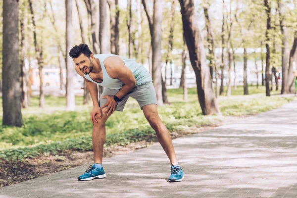 Handsome man in sportswear and sneakers touching injured leg while standing in park — Stock Photo