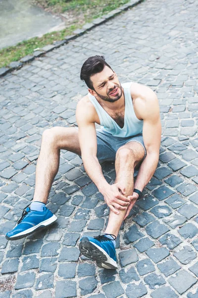 Young sportsman touching injured leg while sitting on pavement and suffering from pain — Stock Photo